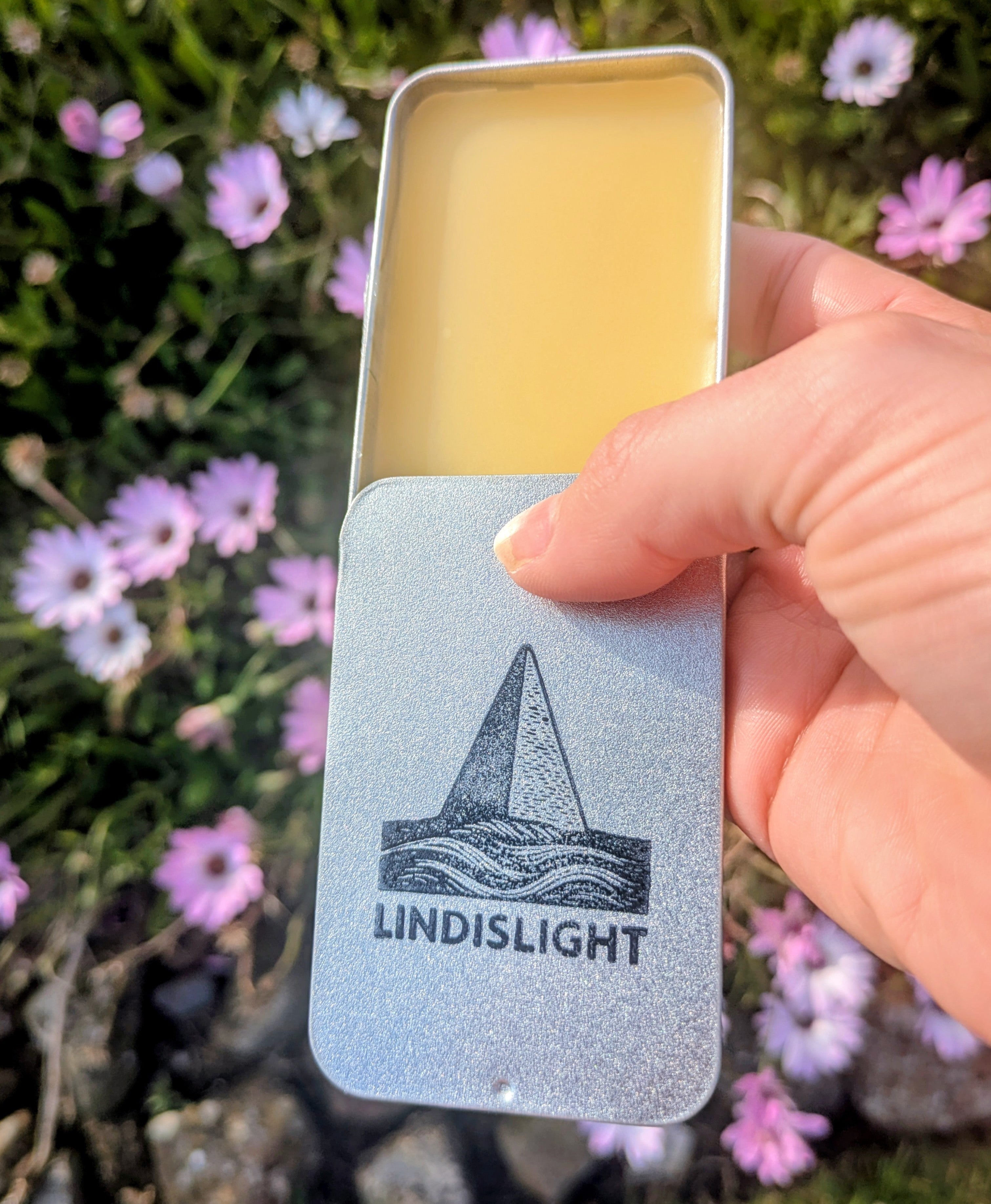 Hand holding open Lindislight balm tin showing full logo and yellow balm, with purple flowers in background